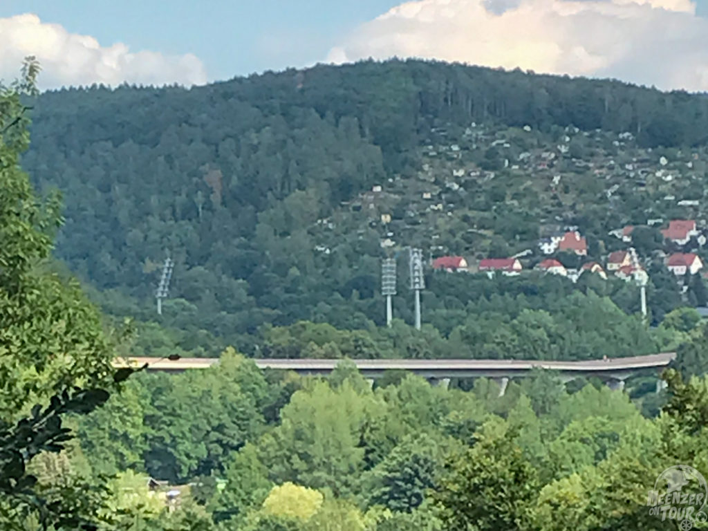 Bewaldeter Hügel mit vier Flutlichtmasten und einem Neubaugebiet und einer Straßenbrücke.