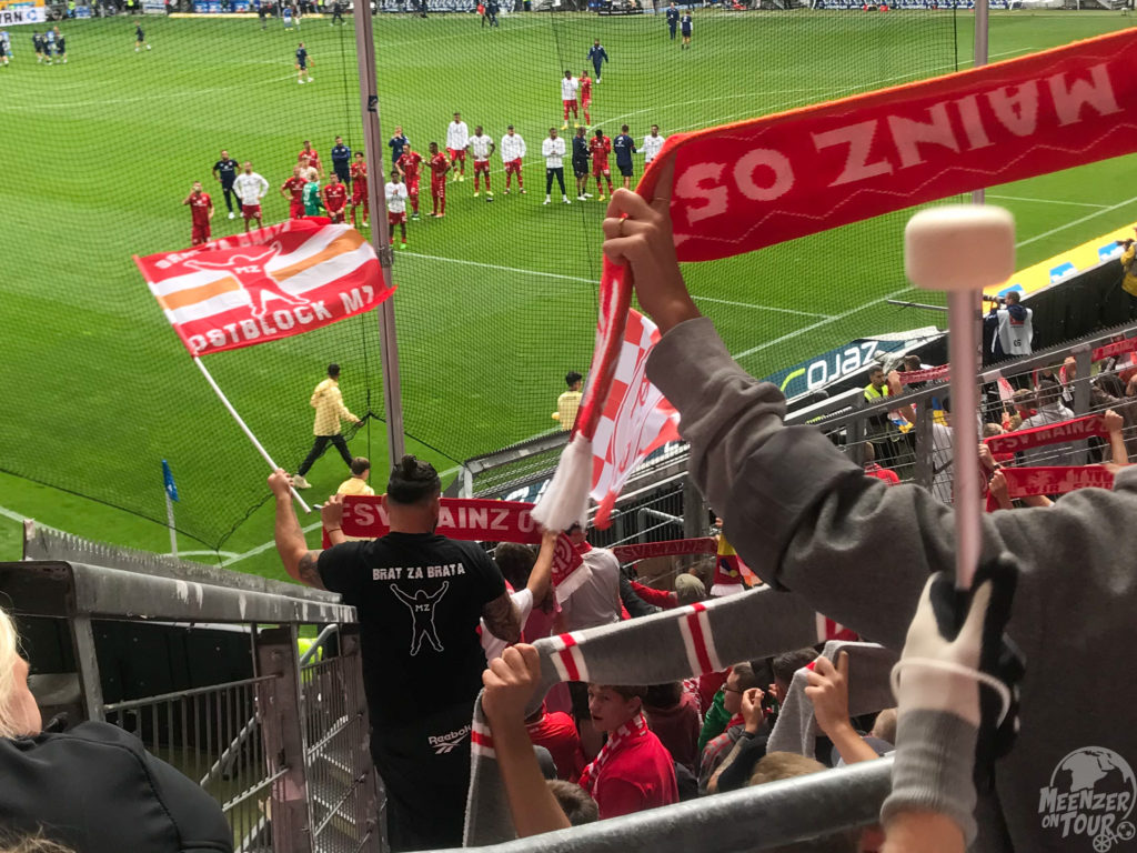 Fans mit Schals und Fahnen und Manschaft in einem Stadion stehen sich gegenüber.